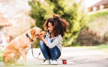 A young girl wearing a striped shirt is sitting outside on a skateboard while petting a large dog on a leash.