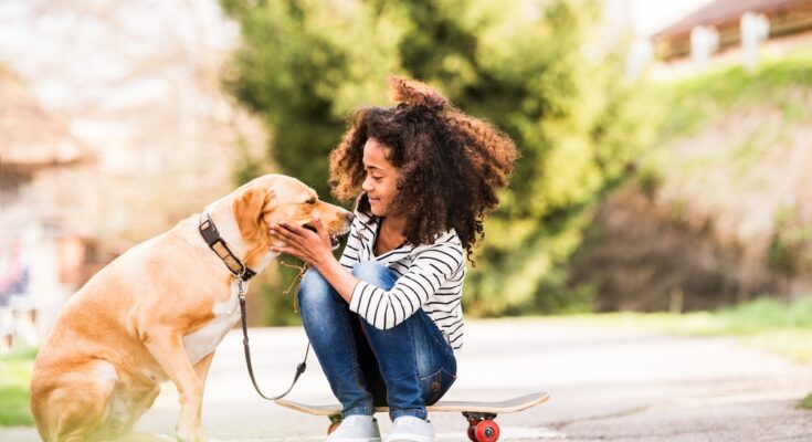 A young girl wearing a striped shirt is sitting outside on a skateboard while petting a large dog on a leash.