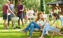 A backyard party. Three men stand around a grill and two women chat in lawn chairs while holding cocktails.