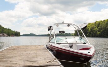 A blue and white boat sits tied up to a wooden dock on a body of water. Trees surround the water in the background.