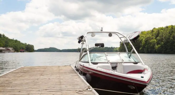 A blue and white boat sits tied up to a wooden dock on a body of water. Trees surround the water in the background.