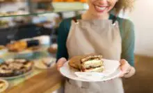 A close-up view shows a sandwich wrapped in brown wax paper and sitting on a white plate. A smiling woman holds the plate.