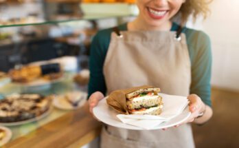 A close-up view shows a sandwich wrapped in brown wax paper and sitting on a white plate. A smiling woman holds the plate.