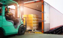 A person drives a green forklift toward a loading dock to load multiple pallets of cargo into a large freight truck.