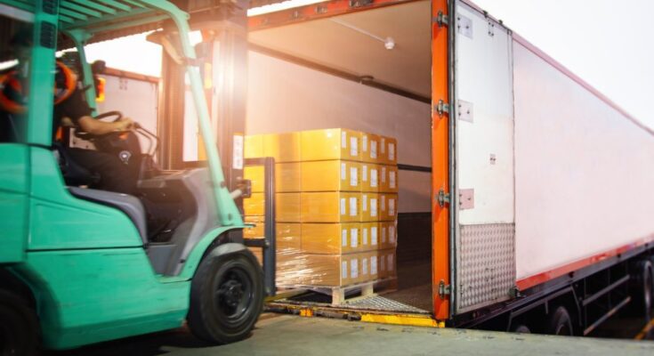 A person drives a green forklift toward a loading dock to load multiple pallets of cargo into a large freight truck.