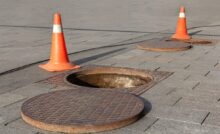 Two orange safety cones appear next to manholes on a sidewalk with the manhole covers right next to them.