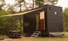 A rectangular tiny home on wheels parked near trees in a forest. An awning covers the entrance and a pair of Adirondack chairs.