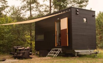 A rectangular tiny home on wheels parked near trees in a forest. An awning covers the entrance and a pair of Adirondack chairs.