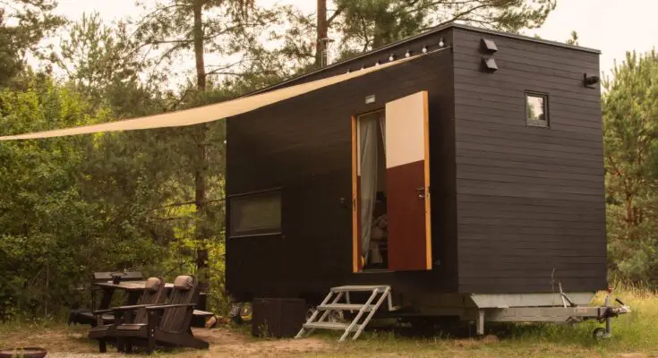 A rectangular tiny home on wheels parked near trees in a forest. An awning covers the entrance and a pair of Adirondack chairs.