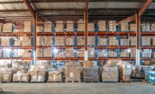 A warehouse contains vertical storage with pallets wrapped in clear plastic. Each pallet contains cardboard boxes.