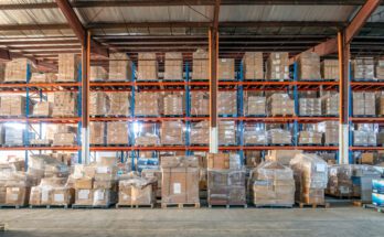 A warehouse contains vertical storage with pallets wrapped in clear plastic. Each pallet contains cardboard boxes.