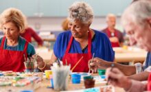 A group of seniors in red aprons sitting at a table, holding paintbrushes while painting on white pieces of paper.