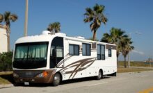 A large white and brown RV parked on the side of a road with palm trees in the background on a sunny day.