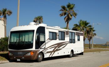 A large white and brown RV parked on the side of a road with palm trees in the background on a sunny day.