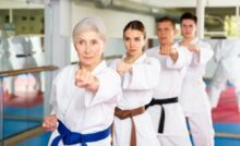 A row of people wearing different colored belts practice karate in a mirrored studio. All have their left fists forward.