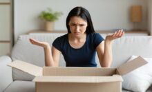 An annoyed woman sits on a couch and lifts her hands up in exasperation as she looks at the contents of a cardboard shipping box.