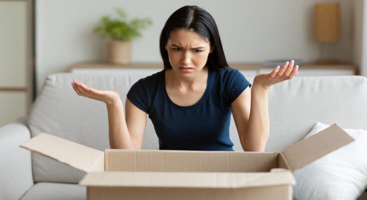 An annoyed woman sits on a couch and lifts her hands up in exasperation as she looks at the contents of a cardboard shipping box.