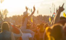 A large crowd of fans with flags at a music festival. A girl sits on a guy's shoulders, her arms in the air.