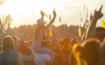 A large crowd of fans with flags at a music festival. A girl sits on a guy's shoulders, her arms in the air.