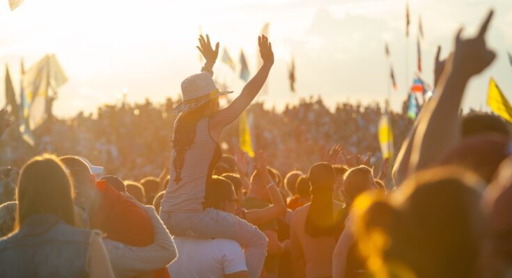 A large crowd of fans with flags at a music festival. A girl sits on a guy's shoulders, her arms in the air.