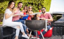 A group of people with white plates and red cups sit next to a grill inside of a truck bed while eating hamburgers.
