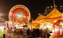 A blurred view of a carnival. There are carnival games to the right and a moving Ferris wheel in the background.