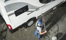 An overhead view of a man in a blue hat and blue overalls using a power washer to clean the exterior of a white RV.