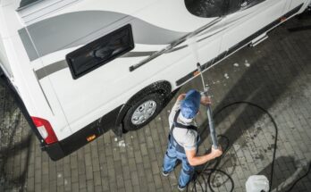 An overhead view of a man in a blue hat and blue overalls using a power washer to clean the exterior of a white RV.
