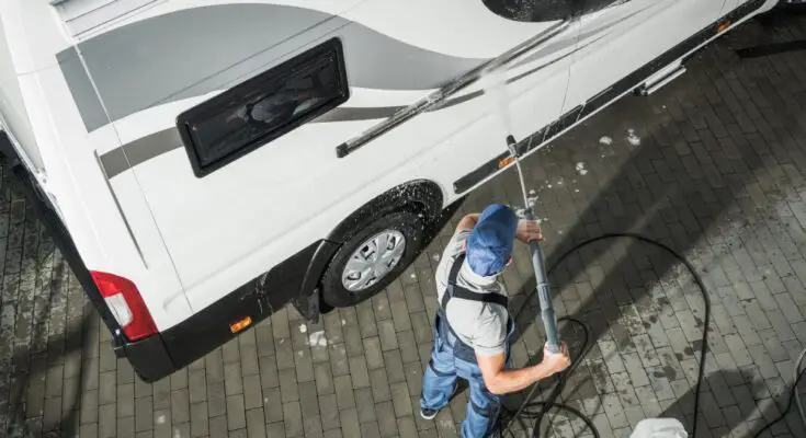 An overhead view of a man in a blue hat and blue overalls using a power washer to clean the exterior of a white RV.