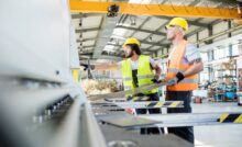 A man in a yellow vest and hardhat and a man in an orange vest and yellow hardhat in a factory.