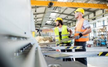 A man in a yellow vest and hardhat and a man in an orange vest and yellow hardhat in a factory.