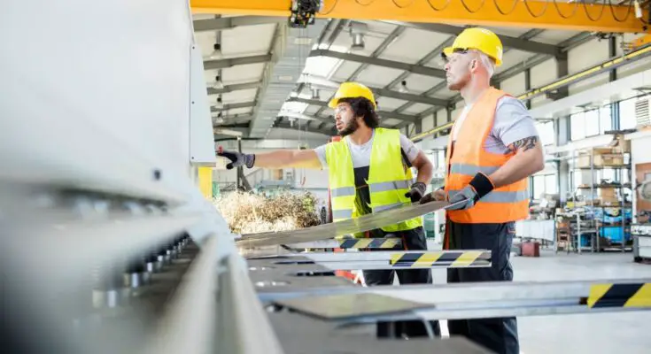 A man in a yellow vest and hardhat and a man in an orange vest and yellow hardhat in a factory.