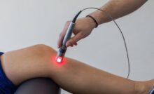 A close-up of a doctor focusing red laser light on a spot just below the bent knee of a patient.