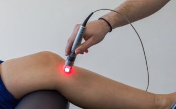 A close-up of a doctor focusing red laser light on a spot just below the bent knee of a patient.