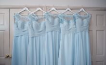 Five light-blue, floor-length bridesmaid dresses hang from white hangers against white doors with gold knobs.