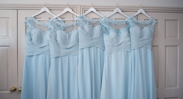 Five light-blue, floor-length bridesmaid dresses hang from white hangers against white doors with gold knobs.