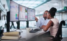 A man with glasses pointing at three monitors showing graphs while sitting next to a coworker wearing a pink shirt.
