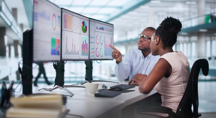 A man with glasses pointing at three monitors showing graphs while sitting next to a coworker wearing a pink shirt.