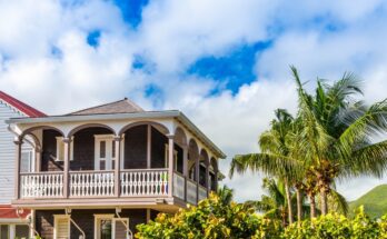 A two-story home with a balcony view next to palm trees basks in warm sunlight. A window on the first floor is open.