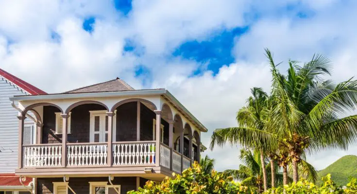 A two-story home with a balcony view next to palm trees basks in warm sunlight. A window on the first floor is open.