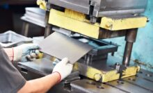 A team member wearing white protective gloves puts a small piece of sheet metal into a yellow sheet metal machine.