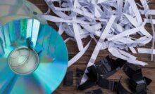 A close-up view shows a partially shredded CD, shredded paper, and shredded black paper on a wooden surface.