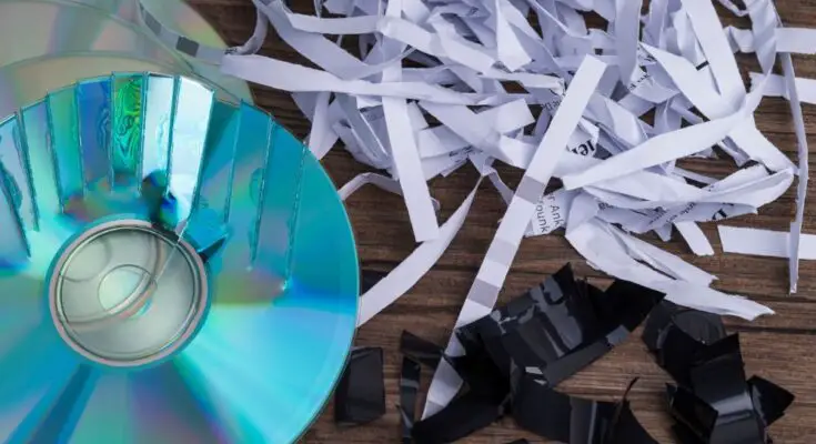 A close-up view shows a partially shredded CD, shredded paper, and shredded black paper on a wooden surface.