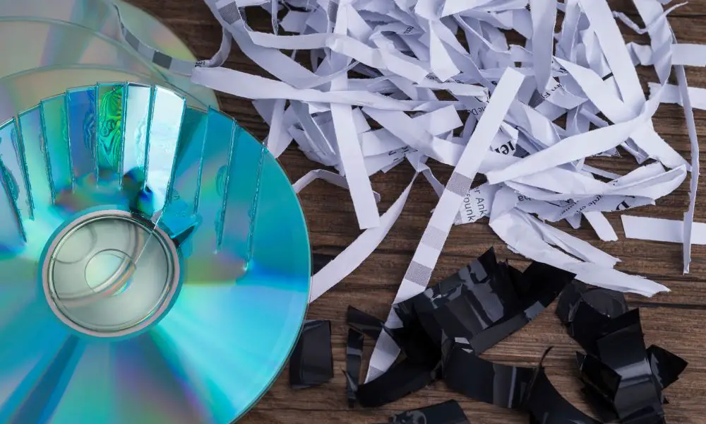 A close-up view shows a partially shredded CD, shredded paper, and shredded black paper on a wooden surface.