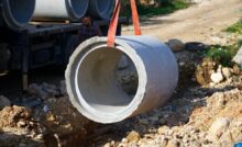 A close-up of a red strap lifting a large cement pipe above a pile of rubble. There is a man standing near the pipe.