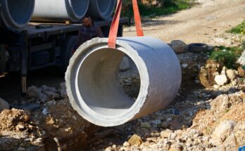 A close-up of a red strap lifting a large cement pipe above a pile of rubble. There is a man standing near the pipe.