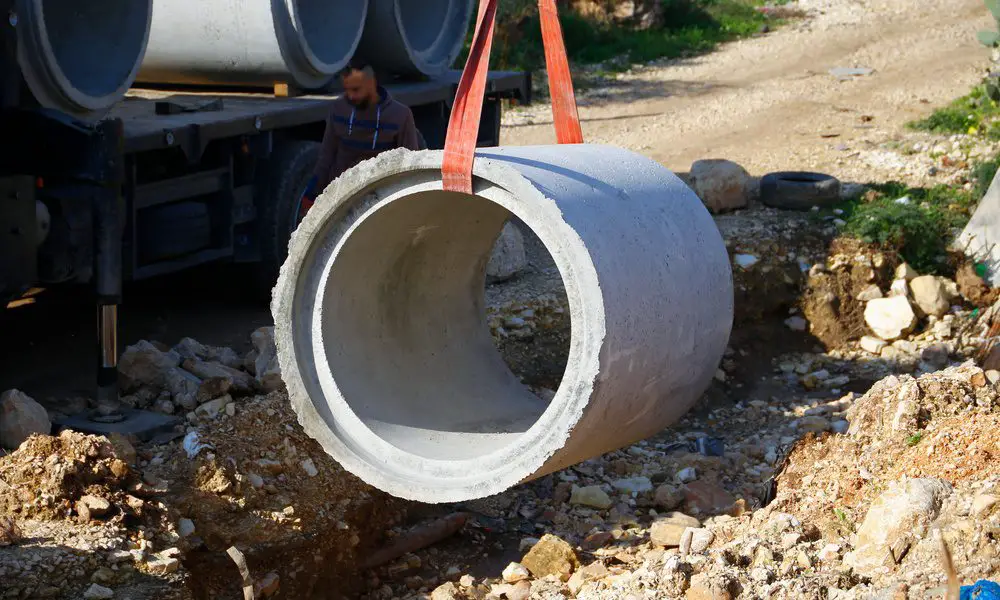 A close-up of a red strap lifting a large cement pipe above a pile of rubble. There is a man standing near the pipe.