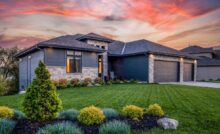 A modern home with gray siding and a brick facade. The lawn is freshly mowed, and the sun is setting in the distance.