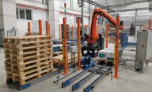 An orange palletizer machine stacks white cardboard boxes on a wooden pallet. A pile of empty pallets is next to the machine.