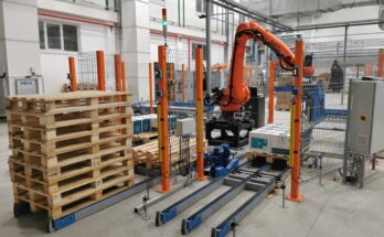 An orange palletizer machine stacks white cardboard boxes on a wooden pallet. A pile of empty pallets is next to the machine.